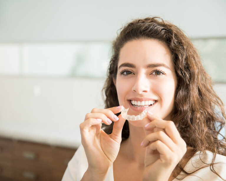 A smiling woman holding a clear dental aligner in front of her mouth at home.