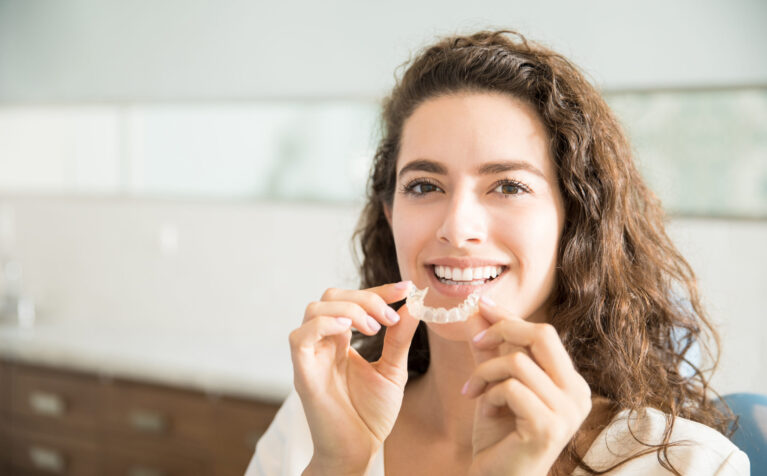A smiling woman holding a clear dental aligner in front of her mouth at home.