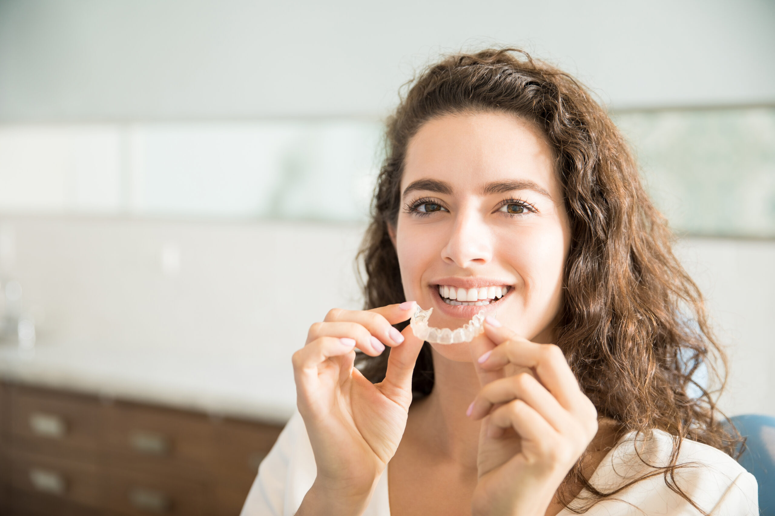 A smiling woman holding a clear dental aligner in front of her mouth at home.