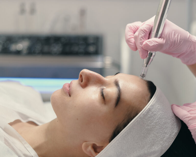 A woman lying on a treatment bed while a clinician wearing gloves performs a microneedling facial treatment with a handheld device