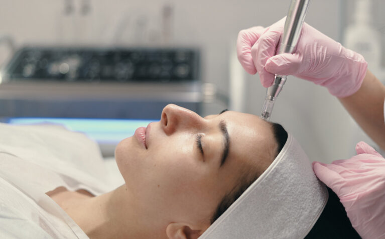 A woman lying on a treatment bed while a clinician wearing gloves performs a microneedling facial treatment with a handheld device